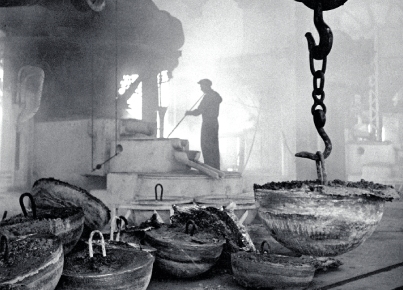 Interno industriale con operaio in controluce e grandi contenitori appesi.