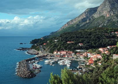 Veduta di un porto costiero con barche, case sul promontorio e montagna sullo sfondo.