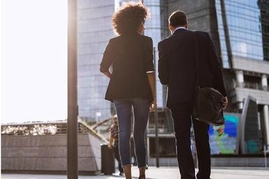 Due persone camminano in un centro direzionale tra grattacieli e passaggi coperti, in una scena urbana al tramonto.