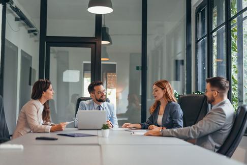 Persone in riunione attorno a un tavolo in ufficio, con un laptop aperto e finestra luminosa sullo sfondo.