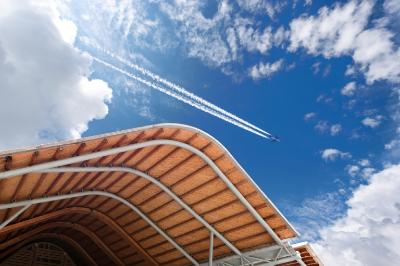 Struttura architettonica con copertura in legno curvata contro un cielo azzurro, con un aereo che lascia una scia bianca