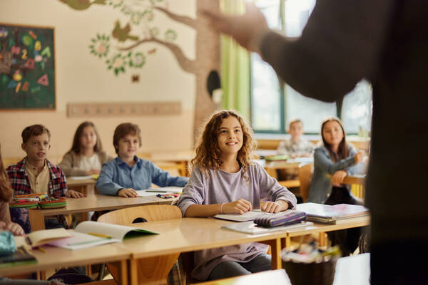 Bambina in classe che sorride seduta al banco mentre studia con i compagni sullo sfondo.
