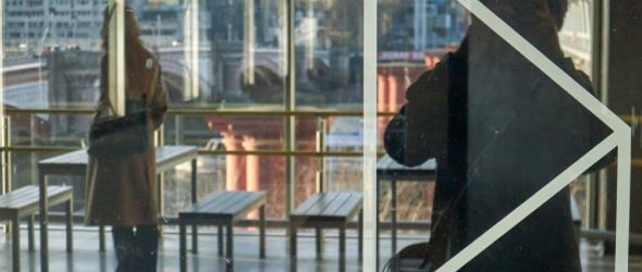 Una persona alza la mano e gesticola, vista attraverso un vetro con riflessi in un edificio urbano.