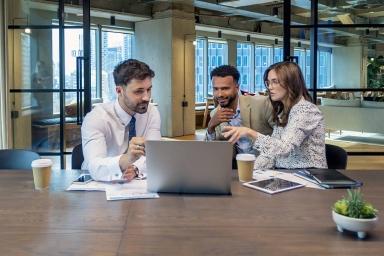 Tre persone lavorano insieme in ufficio, una tiene in mano un bicchiere mentre guardano lo schermo del computer.