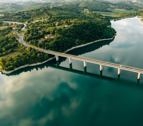 Vista aerea di un fiume con un ponte lungo in mezzo al verde, riflessi nell’acqua e colline sullo sfondo.