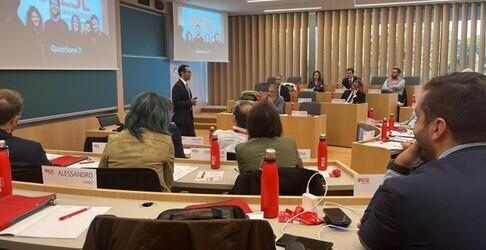 Sessione di conferenza in un’aula: un relatore parla davanti a un gruppo seduto a un tavolo con microfoni e cartelline.