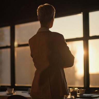 Una persona in piedi davanti a grandi finestre con il sole che tramonta sullo sfondo.