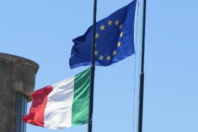 Bandiera dell’Unione Europea e bandiera italiana sventolano davanti a palazzi storici lungo un edificio cittadino.