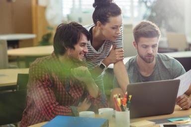 Tre persone sedute a un tavolo che lavorano insieme al computer, con fogli e penne davanti.