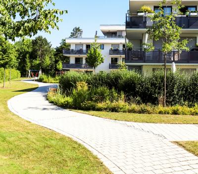 Vialetto curvo in un giardino verde con alberi e aiuole, davanti a edifici residenziali moderni.