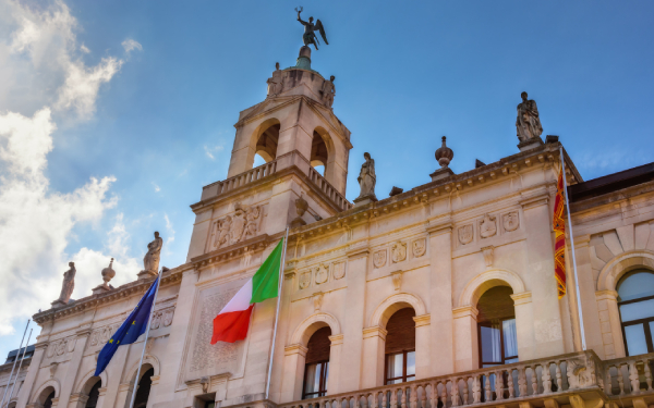 Edificio neoclassico italiano riccamente decorato, con statue, finestre ad arco, torre dell'orologio e bandiere italiana ed europea, sullo sfondo di un cielo azzurro intenso.