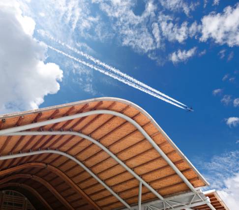 Struttura architettonica con copertura in legno curvata contro un cielo azzurro, con un aereo che lascia una scia bianca.